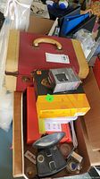 Photo showing vintage Kodak Brownie camera with attached flash reflector on top of a vintage red and beige leather case, surrounded by boxes including Kodak film and accessories.