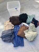 Photo of various folded sweaters, cardigans, a dress, and pants laid out on concrete along with a plastic storage container