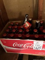 Cardboard box containing multiple unopened classic Coca-Cola bottles with red caps and one dark bottle with cork stopper visible at the top.