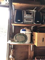 Emerson stereo system with two black speakers placed on a wooden shelf, white enamel crock, decorative green plate, cardboard box, and wooden kitchen utensil beneath.