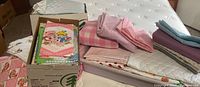 Photo showing two boxes and a tote filled with pastel colored fabric pieces, mainly pink and blue, laid out on a mattress. Includes folded fabrics and loose pieces with visible cartoon character prints.