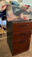 Three-drawer filing cabinet with dark wood-grain finish and plastic bin of crochet blankets on top