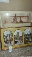 Photo showing three framed pictures stacked against a wall, with the largest featuring three cityscape illustrations with people and architectural elements.