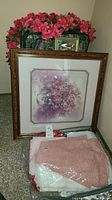 Photo showing one large floral framed picture leaning on the wall with basket of faux pink flowers behind and several pink and white folded fabric tablecloths wrapped in plastic in front.