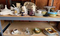 Shelving with assorted vintage Pyrex glass casserole dishes, glass candlesticks, mugs, plates, and serving bowls showing various glassware and ceramic items staged together.