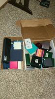 Two open cardboard boxes containing various empty jewelry boxes in multiple colors and sizes placed on carpet.