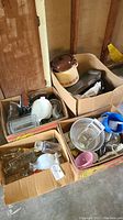 View of three cardboard boxes filled with various kitchen items including a large ceramic pot, glass jars, metal pans, plastic bowls, and other utensils.