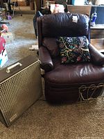 Photo of dark brown leather reclining chair with a decorative floral pillow on the seat and a vintage beige metal fan placed on the floor beside it.