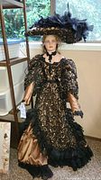 Full view of the porcelain doll standing near a window, showing entire gown, hat, and facial features.