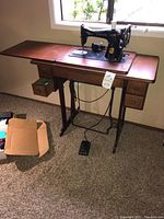Wide view of vintage Singer sewing machine on wooden sewing table with four drawers and metal treadle base. Machine has black body with gold decals. Cardboard box on floor near table.