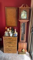 Full view of the vintage wooden grandfather clock next to a wooden chest of drawers with assorted decorative items on top, including porcelain figurines and small collectibles. Clock is tall with a glass door showing pendulum.