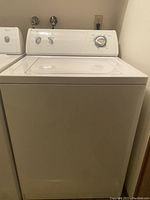 Full front view of the white Whirlpool top load washer showing its lid, control panel with dials, and water hookups behind.