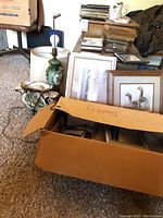 Photo shows two vintage lamps, lamp shades, various framed art and frames partially packed in cardboard box labeled 'frames' set on carpeted floor next to cardboard boxes and furniture.