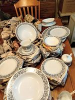 Full view of various pieces wrapped in newspaper on table showing plates, bowls, saucers, gravy boat, sugar bowl and cups with black floral decorative pattern on white dishware