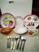 Full view of children's dishware set showing Dora the Explorer plate, Mother Goose bowl, alphabet plates, Strawberry Shortcake mug and utensils.