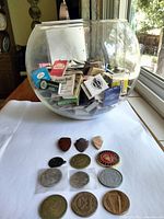 Glass bowl filled with numerous matchbooks; arrowhead-shaped tokens and various coins arranged in front on white surface near window.