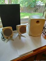 Photo showing two small white Boston Acoustics satellite speakers with stands next to a larger subwoofer speaker on a table with outdoor background.
