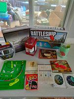 Photo showing game collection including boxed Trivial Pursuit Genus III edition, Battleship electronic edition box, small red mailbox bank, baseball board game, Uno card game, several books, dice and suncatcher items arranged on a table.