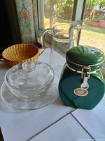 Glass pitcher, glass bowl with lid, green canister with clasp lid, and woven basket on table near window