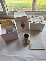 Michael Kors Access smart watch displayed with its box, charging cable, and quick start guide on a table near a window.