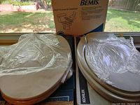 Two toilet seat covers displayed side by side on top of their boxes by a window, one Kohler off white and one Bemis bone color.