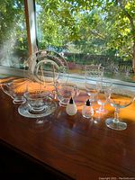 Vintage crystal and glassware set displayed on wooden surface by window, including various glasses, plates, cream and sugar containers, and salt & pepper shakers.