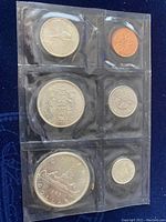 Top view of the six coins including 1955 silver dollar, smaller quarters, dimes, and penny, all in clear plastic packaging within black felt case.
