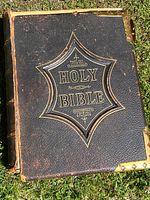 Full view of large black leather Holy Bible with brass corner fittings, showing wear on cover edges and corner. Bible title embossed on front in gold.