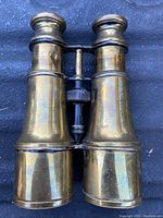 Front view of antique polished brass nautical binoculars on a dark surface. Shows overall shape and condition including natural patina and minor surface blemishes.
