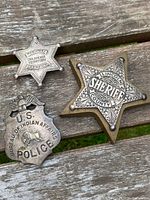 Three antique sheriff badges displayed on a wooden surface showing front details including 'Deputy Oklahoma Territory Marshal', 'U.S. Bureau of Indian Affairs Police', and 'Lincoln County Sheriff' mounted on brass star.