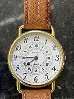 Close-up photo of a round gold-tone Watch-It quartz watch with white dial featuring both Arabic and Roman numerals and brown leather strap