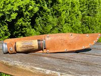 Knife sheathed in a brown leather holder with embossed leaf design, laid on wooden surface outdoors.