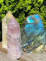 Three polished crystal towers displayed outdoors against green foliage: clear quartz, rose quartz, labradorite