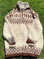 Full front view of a large size Cowichan woven wool sweater featuring brown geometric patterns on cream background, laid flat on grass.