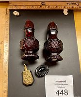 Top view of two dark red glass salt and pepper shakers with detailed textured pattern, small brass topped bottle, and an oval black faceted brooch, ruler showing size