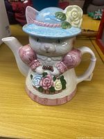 Front-facing photo of the teapot showing the cat figure wearing a blue hat with a yellow rose and pink dress holding a basket of colorful roses.