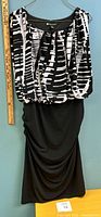 Front view of black skirt with draped detail and black and white patterned top of the dress lying flat against a wall. Ruler for scale shows size approximately 12/14.