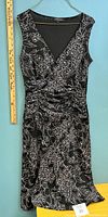 Front view of black sleeveless dress with white floral print hanging on hanger with ruler showing length.