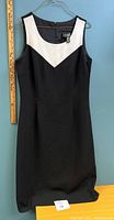 Full view of the sleeveless black dress with white V-shaped neckline panel hanging on a hanger against a blue background with a ruler for size reference.