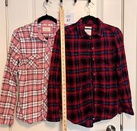 Two flannel tops hanging side-by-side on black hangers, one pink and white plaid and the other red, black and blue plaid. A measuring ruler is positioned between the shirts.