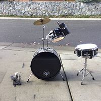 Full view of the kids drum set outdoors, showing bass drum with mounted toms, snare drum, cymbal, and foot pedal separated on the ground.