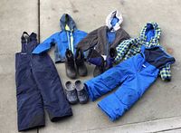 Overview of all items: two rain jackets, two pairs of snow pants, boots, and Crocs arranged on concrete floor.