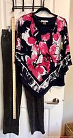 Photo showing full view of dark corduroy pants and flowy floral top hanging on hanger against white door with a measuring ruler beside pants for scale.