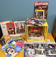 Overview of multiple guitar magazines, instruction books, and guitar tab books laid out on a table with some leaning against a wall.