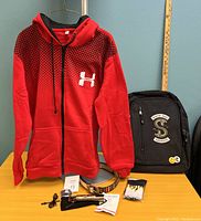 Red and black zip-up hoodie displayed on hanger with black backpack featuring snake emblem, patterned belt, hair clipper, clipper accessories and gloves placed on table underneath hoodie.