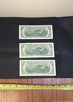 Three consecutive $2 bills laid out horizontally on a dark surface, top sides not shown, with ruler for size reference.