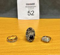 Three rings displayed on a wooden surface: a goldtone ring with sparkle stones, a large oblong stone ring with black onyx, and a silver-tone ring with three opal stones.