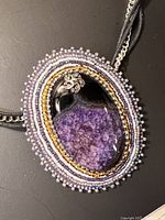 Close-up of the large oval pendant showing the purple semi-precious stone surrounded by detailed beadwork in multiple colors including lavender and gold with a small metallic decorative element.