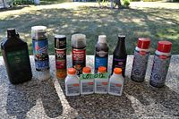 Photo showing a variety of automotive fluids, lubricants, oils, and repair aerosols arranged in a line outdoors on a concrete surface under sunlight.