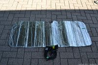 Folded silver reflective automotive sunscreen with bubble-wrap texture laid out on brick pavement, with black laser light and power cord on top.